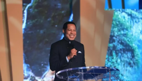 Pastor Chris in a black outfit standing on the stage and holding a microphone