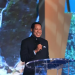 Pastor Chris in a black outfit standing on the stage and holding a microphone